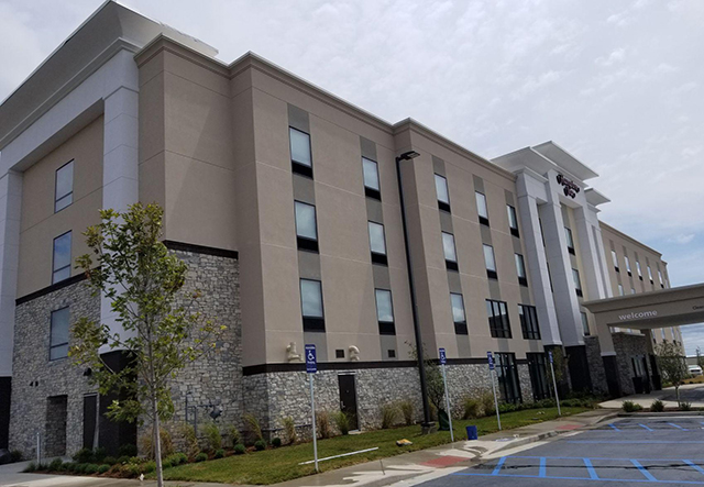 A modern hotel building with four floors, featuring a mix of tan and stone exteriors. The front of the building has an overhang labeled "welcome" near the entrance. There are trees and landscaped greenery around, as well as marked handicap parking spaces in the foreground.