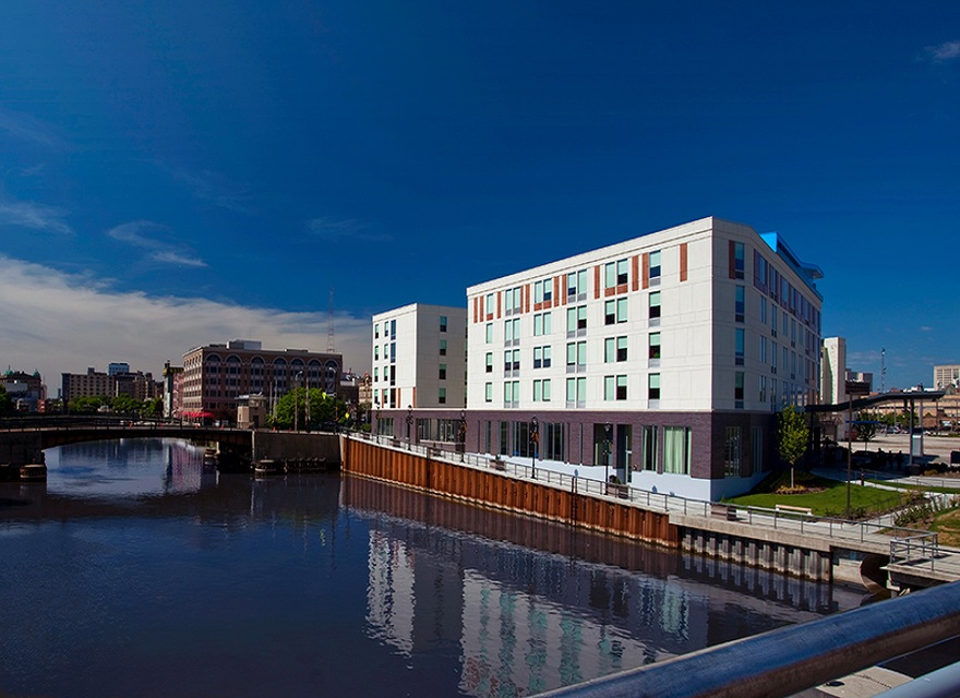 A modern riverside building with large windows stands against a clear blue sky on the right. A bridge and an older building are visible on the left, with the calm river reflecting the structures. A metal railing is in the foreground.
