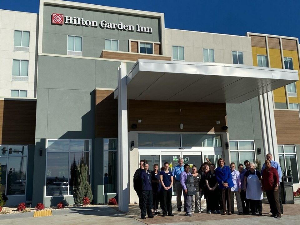 A group of people is standing in front of the entrance to a Hilton Garden Inn hotel. The building behind them has the hotel's sign prominently displayed. The group includes men and women of varying ages, all dressed in business casual and work uniforms.