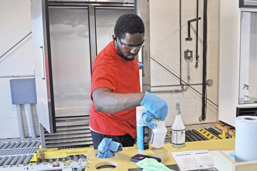 A person wearing a red shirt and protective gloves works at a table with various cleaning supplies and tools. They are in an industrial setting, possibly prepping or cleaning equipment. Safety goggles are worn as part of protective gear.