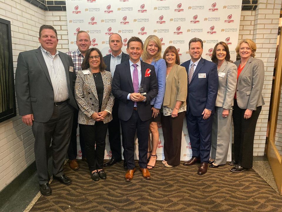 A group of ten people, dressed in business attire, stand together smiling in front of a white backdrop with a red and gray logo. They are positioned in two rows, with five in the front and five in the back. The setting appears to be a formal event.