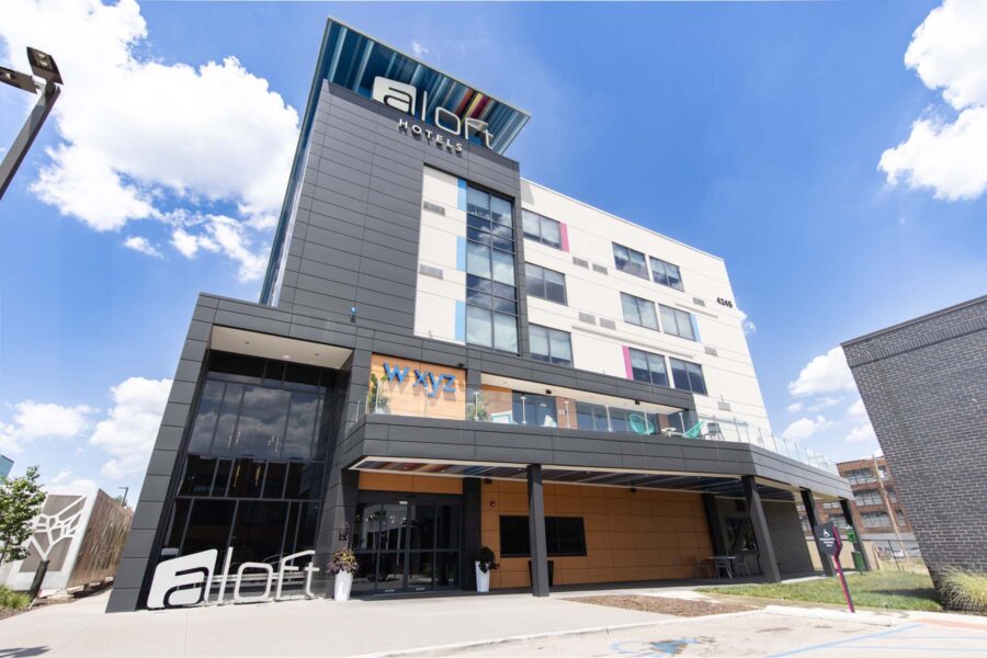 A modern four-story building of Aloft Hotels with tall glass windows, colorful accents, and a prominent sign at the top under a blue sky. The entrance features an outdoor seating area and a covered porch. The ground has a smooth pavement.