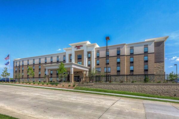 A modern, four-story hotel with a combination of light and dark brown exterior walls, featuring an entrance canopy. American flags are flying near the entrance. A neatly landscaped area with trees and greenery surrounds the building under a clear blue sky.