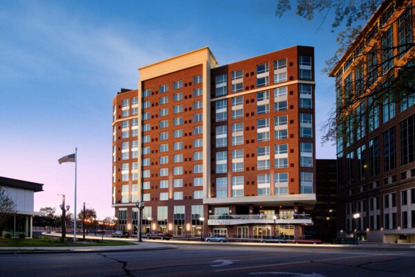 A view of a modern multi-story brick building at dusk, with large windows and balconies. The structure is surrounded by streets, streetlights, and a flagpole. The sky is slightly lit, indicating early evening hours. Cars are parked along the street.