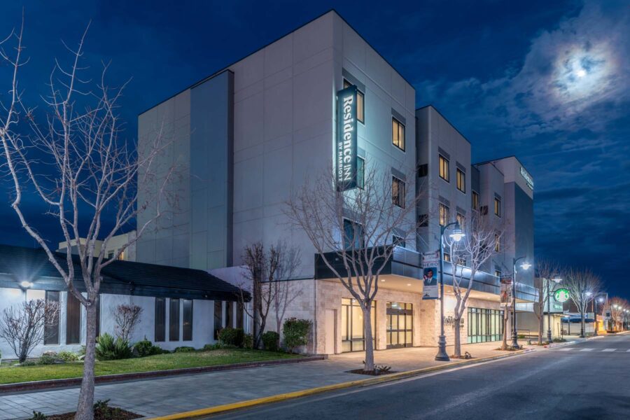 Night view of an illuminated modern building with a Residence Inn by Marriott sign. The building is surrounded by bare trees and lit streetlamps on a quiet sidewalk under a cloudy sky with the moon visible.