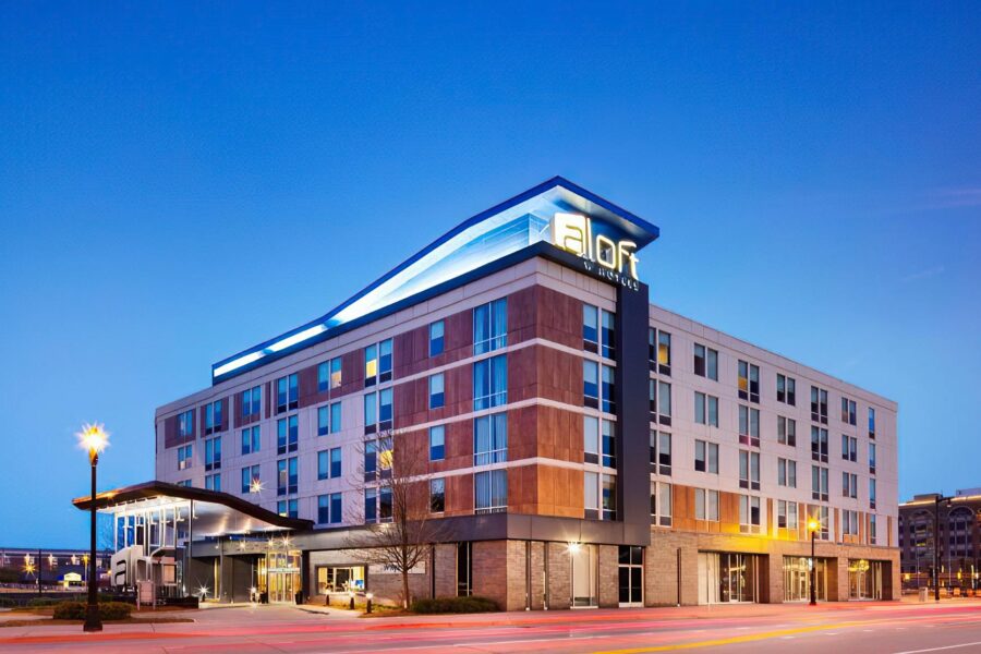 A modern, multi-story hotel building with a sleek design, labeled "Aloft," illuminated at twilight. The façade is a mix of glass and different shades of brown panels. Surrounding the hotel are well-lit streets with light trails from passing cars.