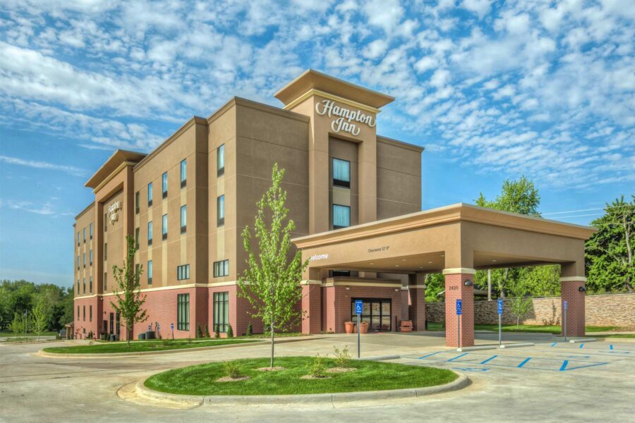 A four-story Hampton Inn hotel building with a beige and reddish-brown exterior is pictured on a sunny day with a partly cloudy sky. The entrance has a covered driveway. The parking lot includes accessible parking spaces, and young trees are planted near the entrance.