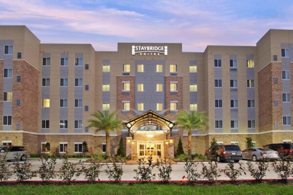 The front view of a Staybridge Suites hotel building at dusk, featuring rows of windows, a well-lit entrance with an archway, and two palm trees on either side of the entryway.