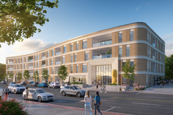 Modern four-story building with large windows, brick and white facade. Green trees and parked cars line the street, while people walk and cycle along the sidewalk. The building features balconies and a central entrance with contemporary design. Clear blue sky.