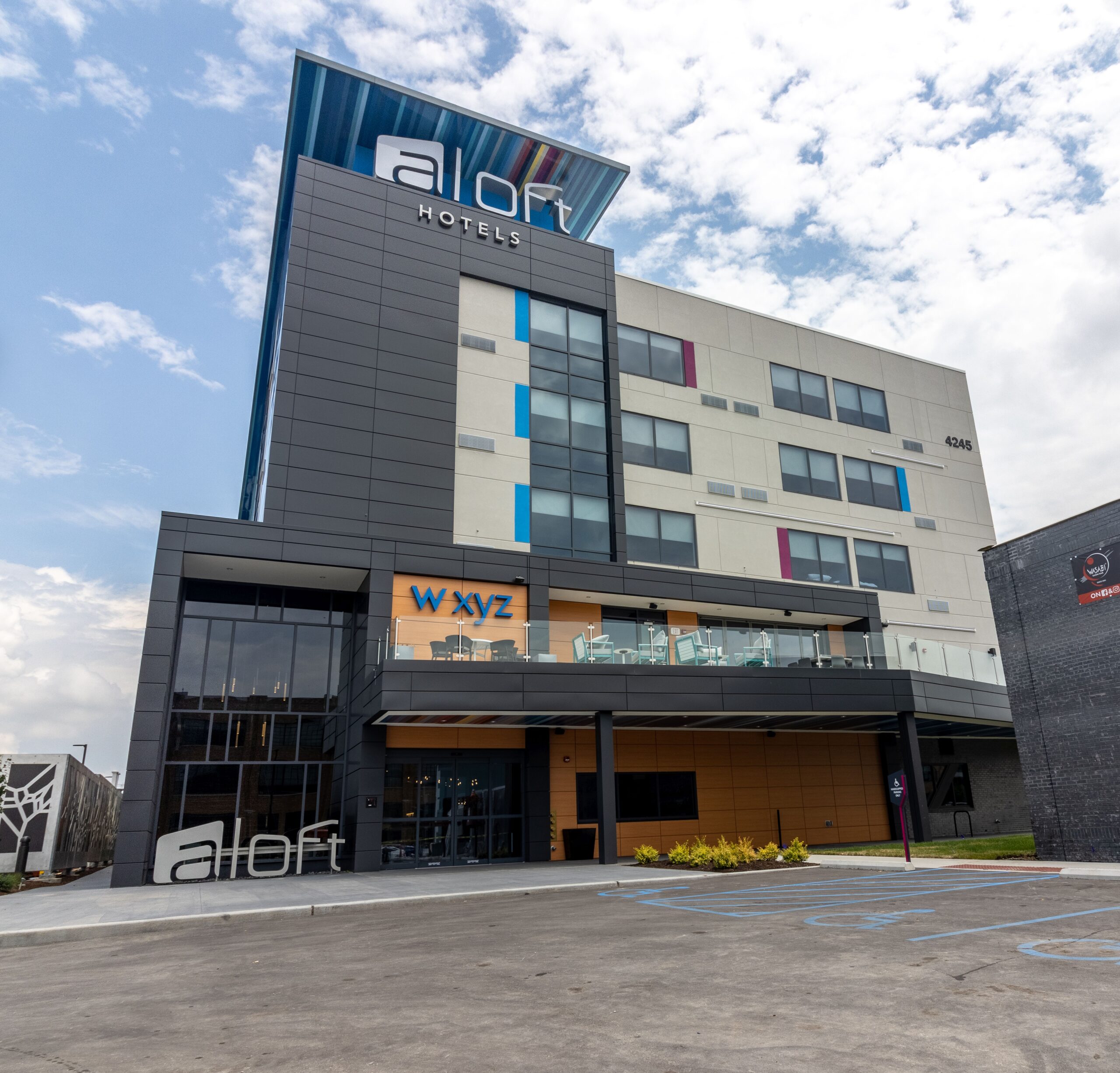 Exterior view of an Aloft hotel building with a modern design, featuring a mix of grey, white, and colorful paneling. The hotel has a prominent sign at the top and a large "aloft" sign near the entrance. A blue and white sky with clouds is visible in the background.