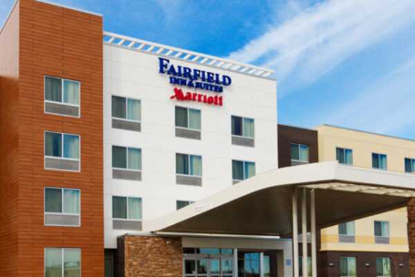 A modern hotel exterior with a mix of white and brown paneling and a covered entrance. The signage reads “Fairfield Inn & Suites Marriott.” There are multiple windows on its four-story structure under a bright blue sky with wispy clouds.