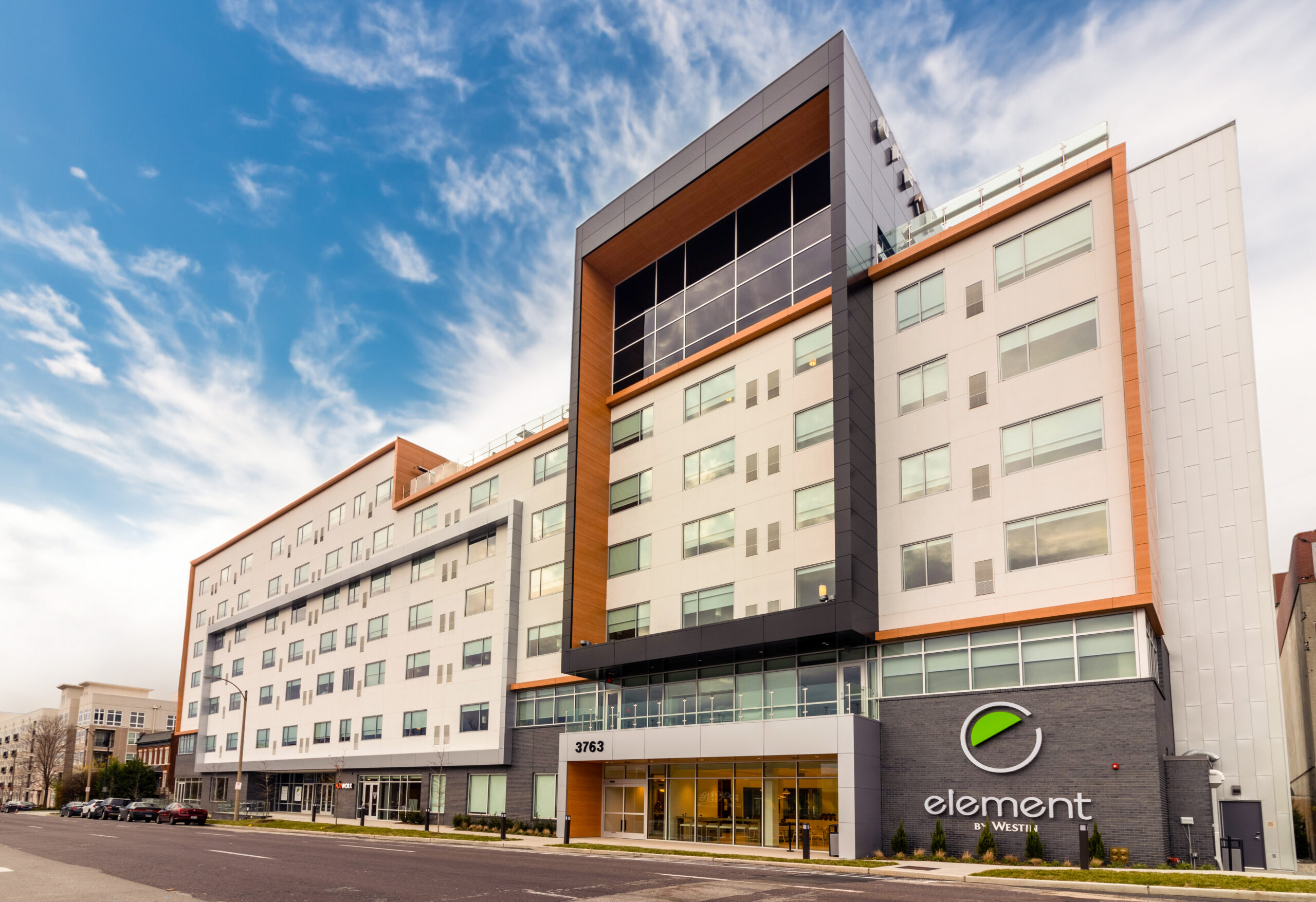 A modern multi-story hotel with the sign "Element by Westin" displayed prominently. The building has a contemporary design with large windows and a mix of white, grey, and wood accents. The entrance features glass doors, and the sky is partly cloudy.