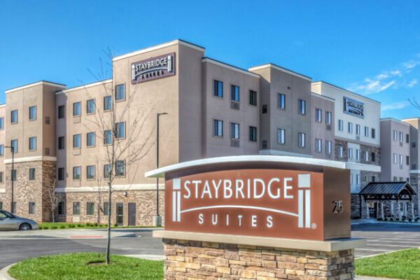 A modern four-story Staybridge Suites hotel building with a stone and tan exterior is pictured on a sunny day. A large, brown sign with "Staybridge Suites" in white letters stands prominently in front of the building, surrounded by manicured grass.