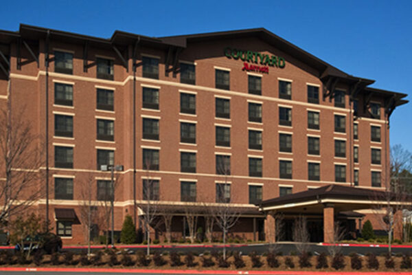 A six-story Courtyard by Marriott hotel with a red brick facade and dark brown trim. The building features multiple windows, a central entrance with a covered drop-off area, and well-maintained landscaping. The foreground includes a partially visible parking lot.