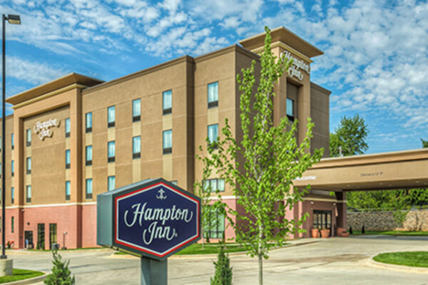 A daylight view of a Hampton Inn hotel, featuring a four-story building with rectangular windows. The hotel sign is prominently displayed in front of the building, which is surrounded by well-maintained landscaping and greenery, under a partly cloudy sky.