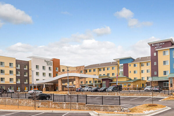 A wide-angle view of a modern hotel complex with multiple connected buildings in various pastel colors. Several cars are parked in the front parking lot, and the sky above is partly cloudy. The hotel's entrance is covered by an archway structure.
