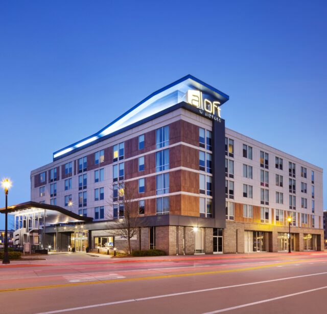 A modern mid-rise hotel building with the illuminated sign "Aloft" on the roof against a dusk sky. The structure features large windows and a combination of brick and panel exterior finishes. A covered entrance leads to the lobby; streetlights brighten the foreground.