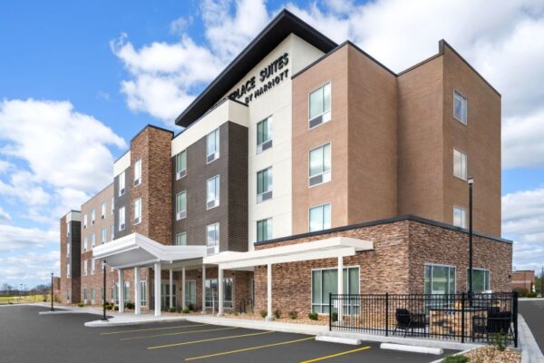 A modern hotel building with four stories, featuring a mix of brick and beige exterior, large windows, and a prominent sign reading "Residence Suites by Marriott." The entrance has a covered canopy, and the surrounding area includes landscaping and a parking lot.