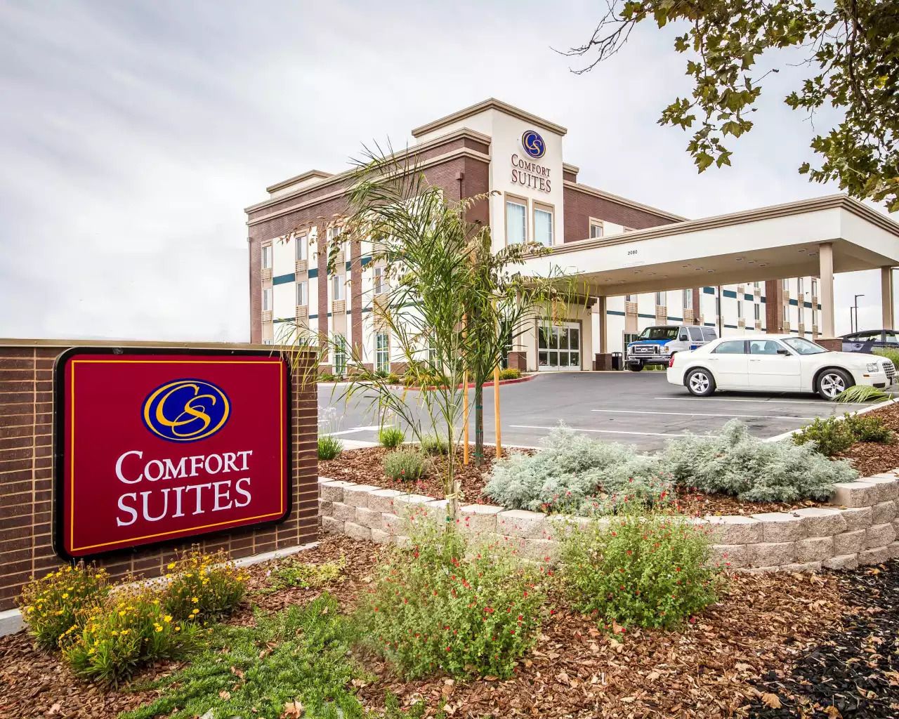 The image shows a Comfort Suites hotel building with a modern facade, featuring large windows and an entrance canopy. A red and blue sign with the Comfort Suites logo stands prominently in front, surrounded by landscaped greenery and a car parked in the driveway.