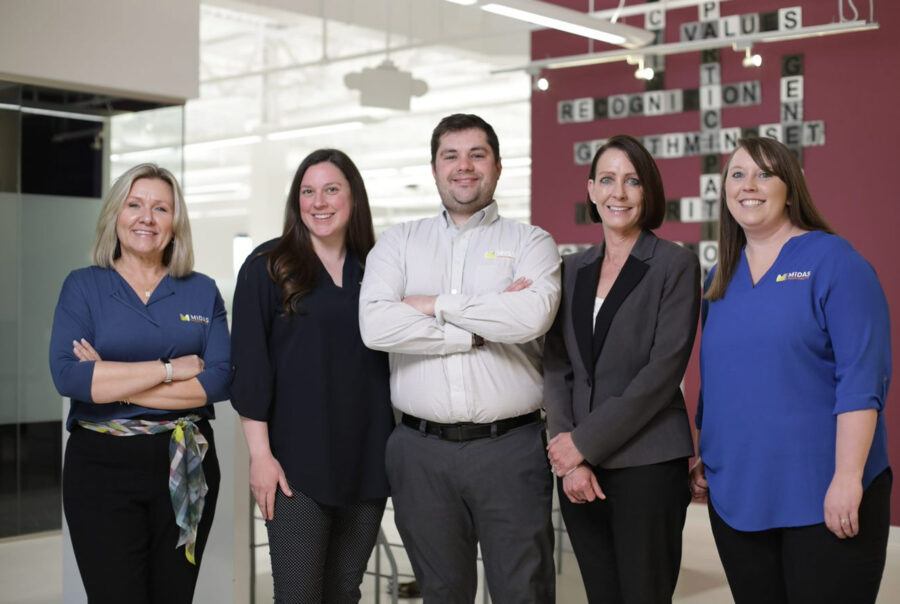 A group of five people standing and smiling in an office setting. Three women are on the left, one man in the middle with arms crossed, and another woman on the right. They are wearing business casual attire and some display company logos. Join us and be part of this vibrant team!