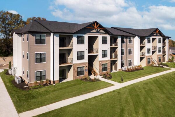 A three-story apartment building with beige and white exterior, featuring wooden accents above the balconies. The building is surrounded by well-maintained lawns and a paved pathway. The sky is blue with scattered clouds.