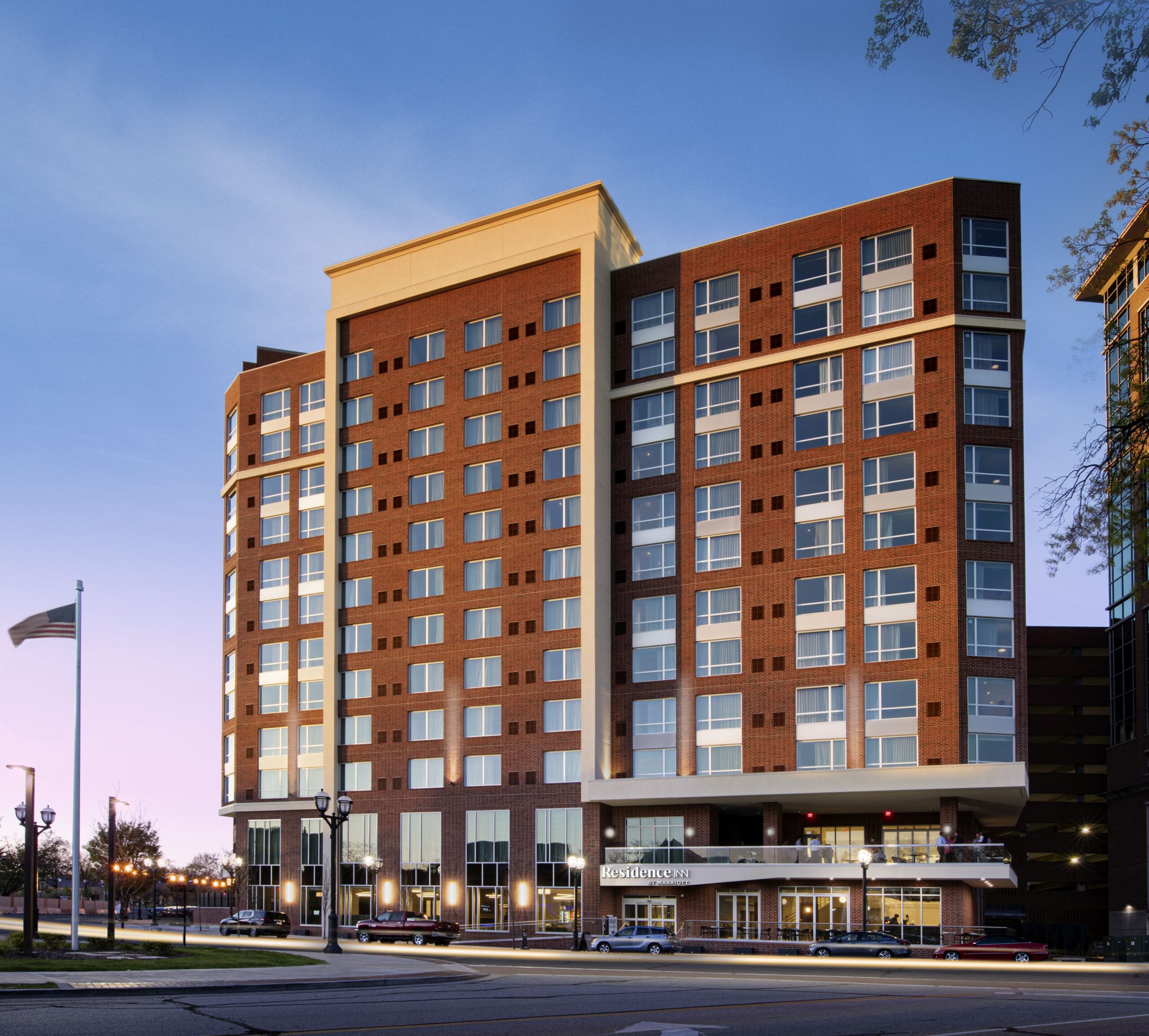 Residence Inn Clayton hotel building with large windows, featuring a prominent entrance with a canopy. The building is well-lit, and the sky is transitioning from dusk to evening. A flagpole with a raised flag is visible in the foreground.